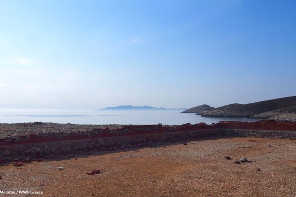 Προαύλιος χώρος και ημιτελής κατασκευή επέκτασης με τη Σύρο να δεσπόζει στο τοπίο, υπενθυμίζοντας τη μετάβαση στη Γυάρο